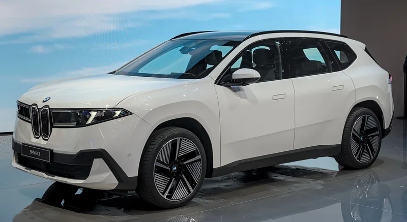 A white BMW iX3, the Neue Klasse electric SUV, seen from the front-left three-quarter angle on an indoor show stand at the IAA Munich auto show in September 2025.