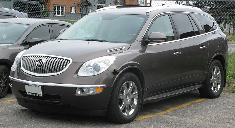 A dark brown 2008 Buick Enclave CXL parked in a lot, seen from the front and its left/driver side, facing image-left, with a chain-link fence behind it.