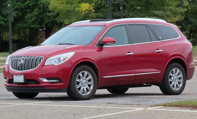 A red 2014 Buick Enclave Leather Group parked on a paved lot with trees behind it, seen from the front and its left/driver side, facing image-left.