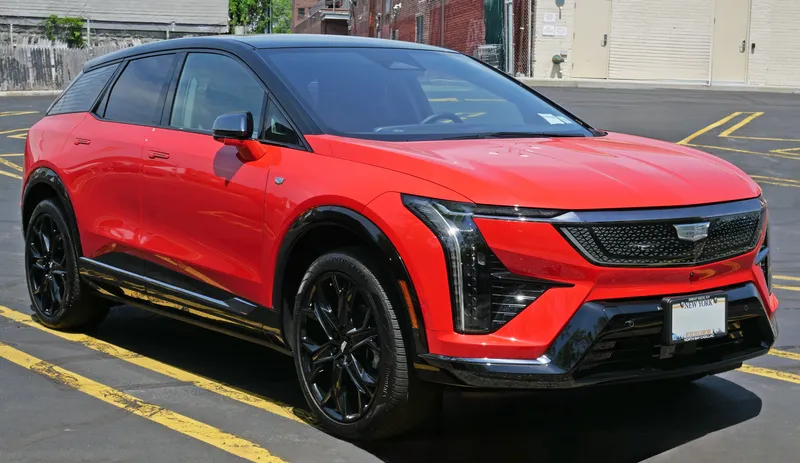 A red 2025 Cadillac Optiq with a black roof and black wheels, seen from the front right, parked in a parking lot. This is a 2025 model, a year RideTruth's catalog does not carry.