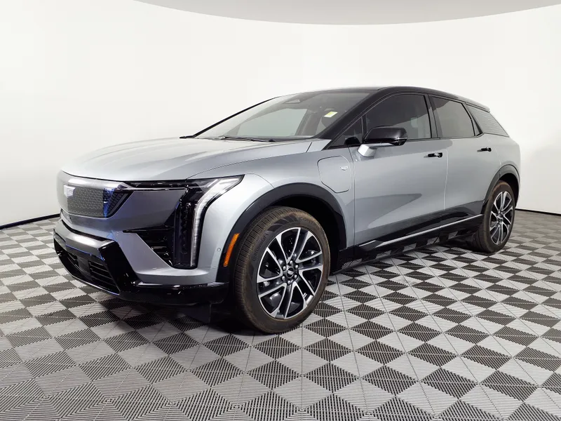 A silver-gray 2026 Cadillac Optiq Sport with a black roof, seen from the front left, parked on a checkered floor in a white showroom.