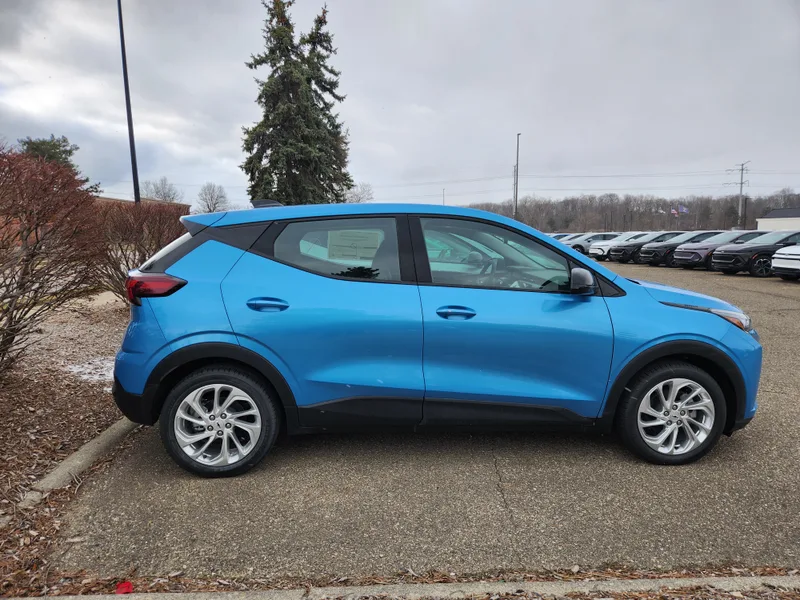 A blue 2027 Chevrolet Bolt, seen from the passenger side, parked in a dealer storage lot with other cars visible in the background.