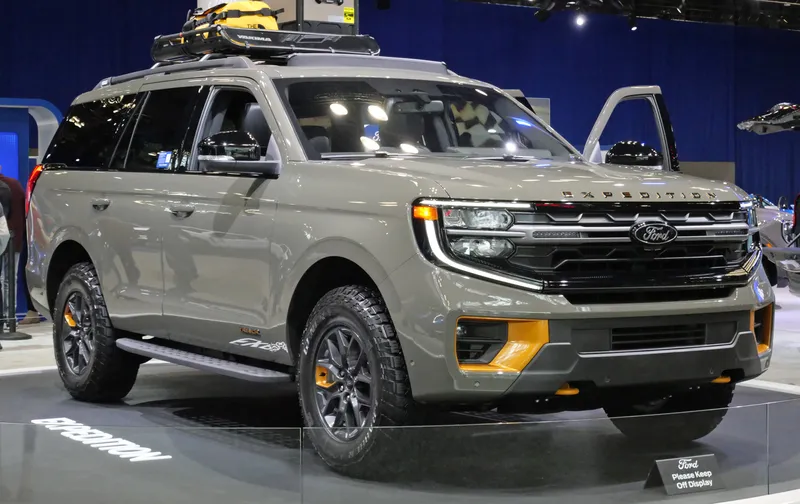 A gray-green 2025 Ford Expedition Tremor with a roof rack on a show-floor display, seen from the front right three-quarter angle, its right headlight and grille facing the camera.