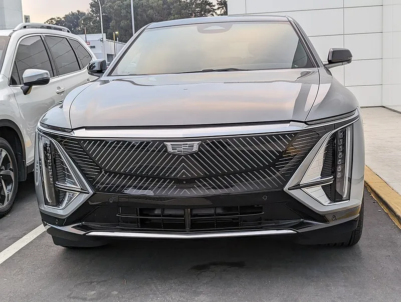 A silver Cadillac Lyriq seen head-on from the front, with its diagonal-patterned grille and vertical headlights, parked outside a dealership in South San Francisco, California. Photographed in August 2023; the model year is not stated.