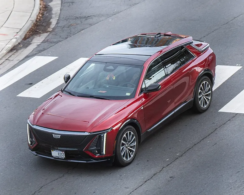 A red Cadillac Lyriq Sport seen from above and the front, showing its left (driver) side as it crosses a painted crosswalk in the Washington, DC area. Photographed in December 2024; the model year is not stated.