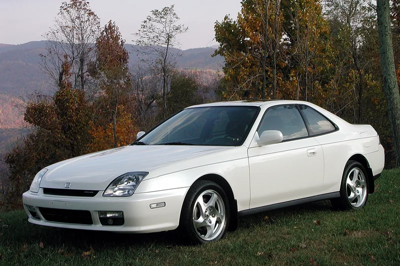 A white 1999 Honda Prelude, seen from the front-left three-quarter angle, parked on grass with autumn trees and hills behind it.