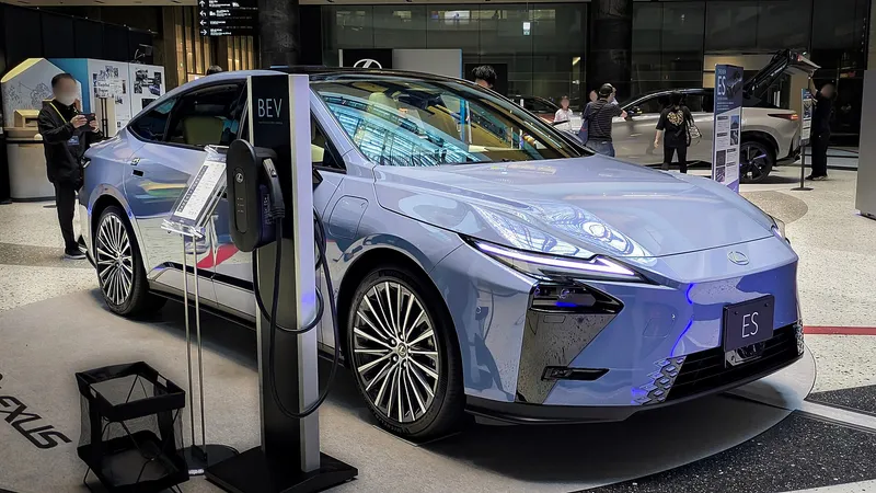 The eighth-generation Lexus ES 350e, front three-quarter view, in a pale silver-blue metallic finish, shown next to a BEV charging post at a Lexus display in Osaka, Japan, May 2026.