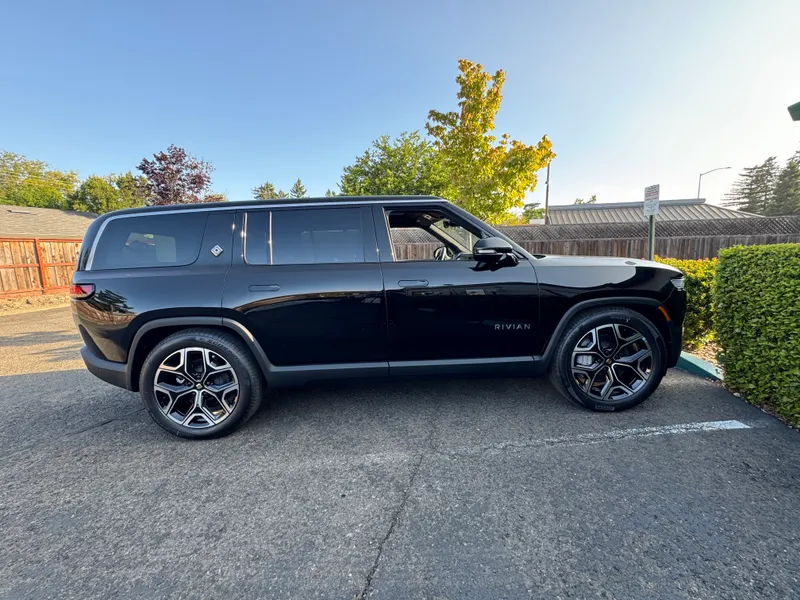 A black Rivian R1S seen from its right side, facing right, parked beside a hedge on a sunny day. The photo is from June 2025 and gives no model year.