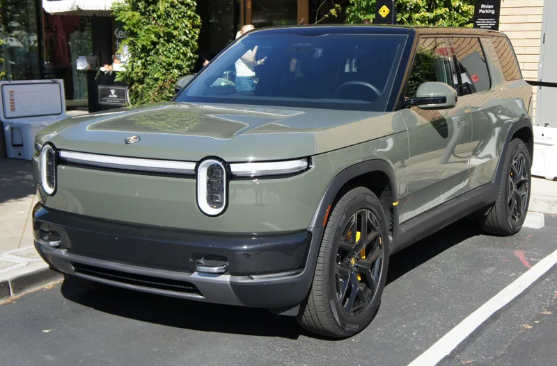 An olive-green Rivian R2 seen from the front left, parked in a street bay beside a curb, with a Rivian parking sign behind it. The photo is from May 2026 and gives no model year.