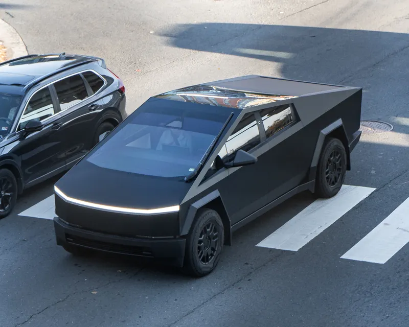 A matte black Tesla Cybertruck, seen from above at a front-left angle, crossing a marked crosswalk next to a dark SUV.