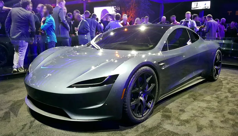 A light silver-gray next-generation Tesla Roadster prototype, seen from the front left, on display indoors at Tesla's November 2017 launch event with onlookers in the background.