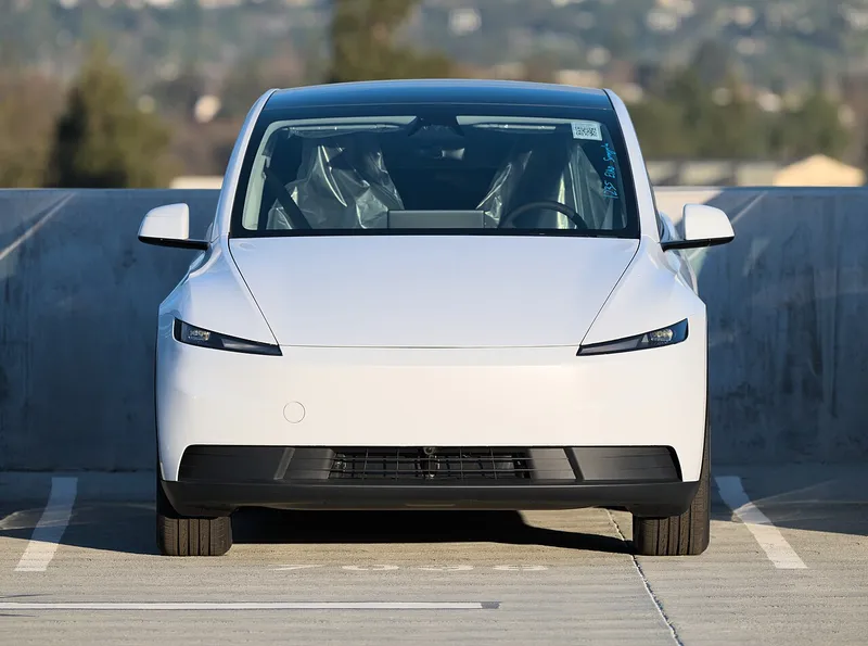 A white Tesla Model Y Standard seen straight on from the front, parked on the top level of a parking structure, with protective plastic still covering the front seats. Commons dates the photo to 2025; it is not a 2026 car or a robotaxi.