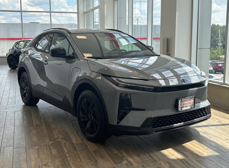 A gray 2026 Toyota C-HR SE AWD, seen from the front right, with a charge-port door visible on the front right fender, on a dealership showroom floor.