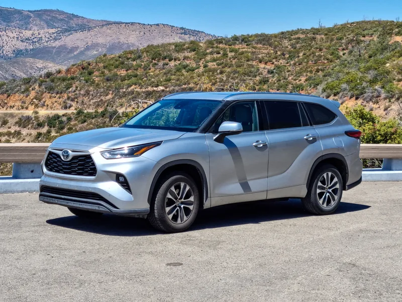 A silver 2022 Toyota Highlander, fourth generation, photographed from the front three-quarter angle, parked at a highway overlook with mountains behind it. This is the outgoing gas-and-hybrid generation Toyota is replacing with the battery-electric 2027 Highlander.