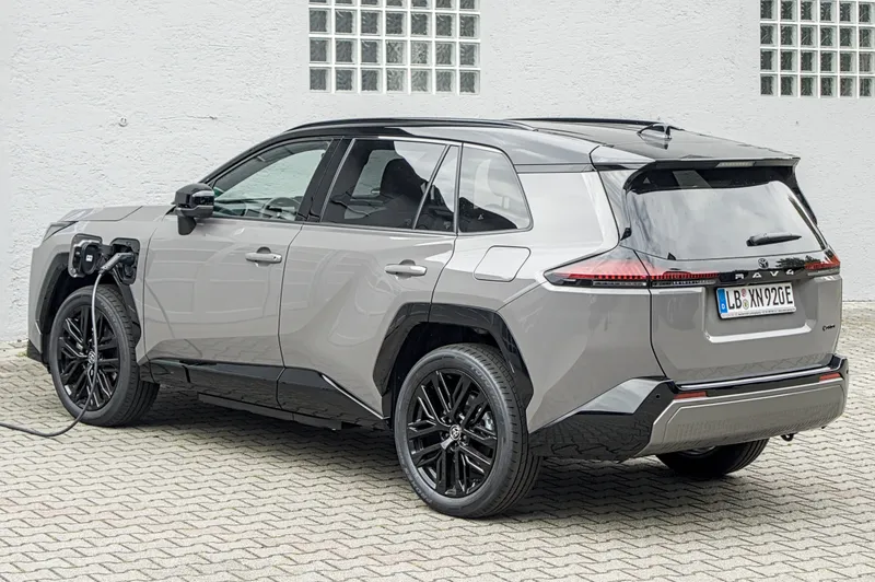 A gray Toyota RAV4 Plug-in Hybrid with a black roof, seen from the rear left (driver's side) at a three-quarter angle, with a charging cable plugged into the front left fender. The car has German license plates and was photographed in Germany in August 2026, so it is a European-market car, not a U.S. one; its model year is not stated.